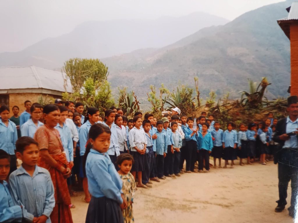 Nepali schoolchildren in blue uniforms lined up outdoors in a mountain village, with a teacher supervising