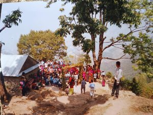 Old photo of a village gathering on a mountainside in Nepal, a crowd of locals near a small building and a floral archway
